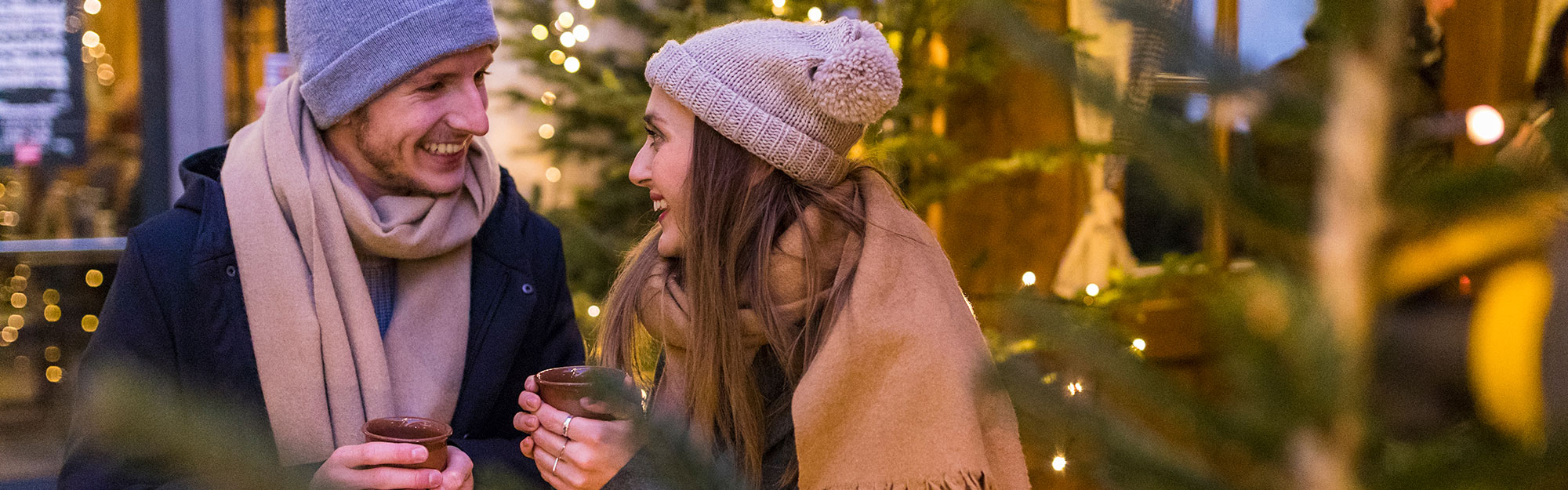 Ett vinterklätt par står utomhus, håller i kaffemuggar och ler mot varandra
